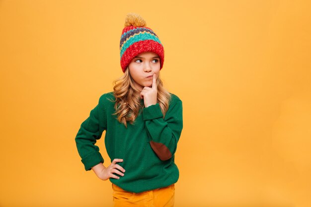 Chica joven pensativa en suéter y sombrero con el brazo en la cadera mirando a otro lado sobre naranja