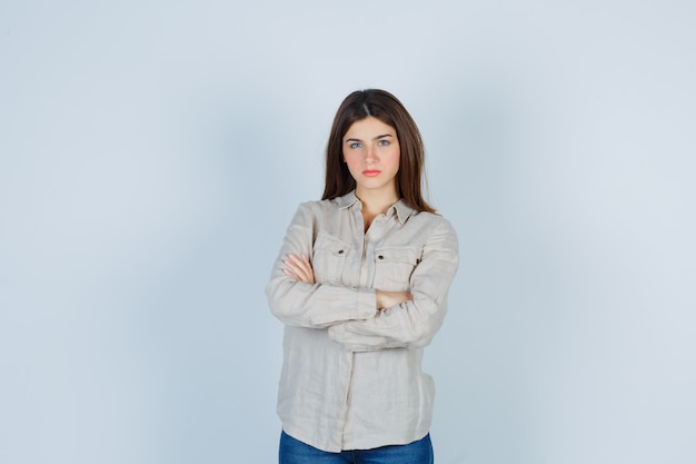 Chica joven de pie con los brazos cruzados en camisa beige, jeans y mirando serio, vista frontal.