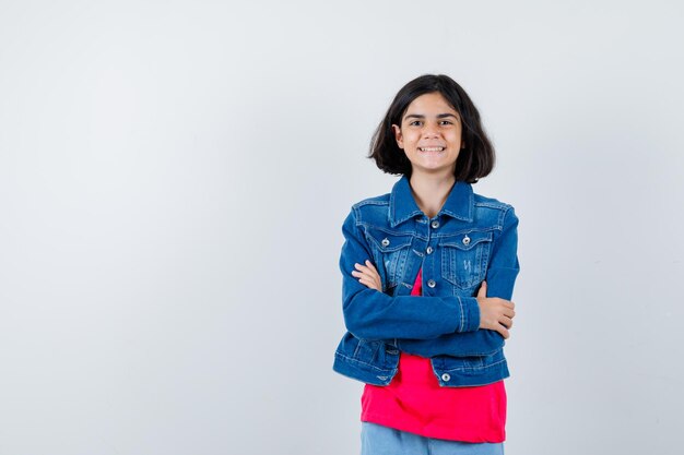 Chica joven de pie con los brazos cruzados en camiseta roja y chaqueta de mezclilla y mirando feliz. vista frontal.