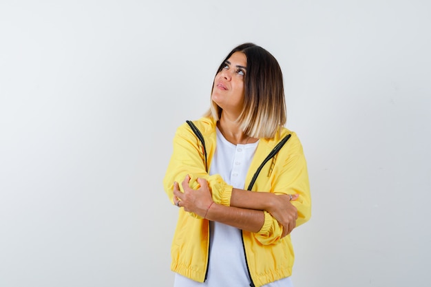 Foto gratuita chica joven de pie con los brazos cruzados, mirando a otro lado en camiseta blanca, chaqueta amarilla y mirando alegre, vista frontal.