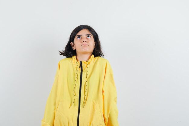 Chica joven de pie con la espalda recta, mirando hacia arriba con chaqueta de bombardero amarilla y mirando seria.