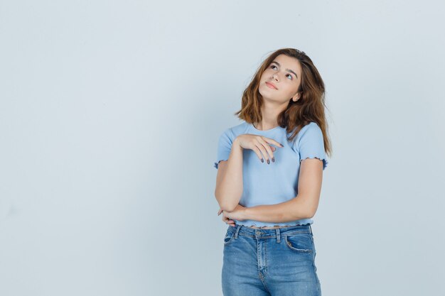 Chica joven de pie en pose de ensueño en camiseta, jeans y mirando linda, vista frontal.