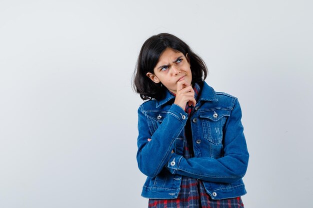 Chica joven de pie en pose de pensamiento en camisa a cuadros y chaqueta de mezclilla y mirando pensativo. vista frontal.