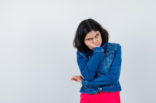 Chica joven de pie en pose de pensamiento en camiseta roja y chaqueta de jean y mirando pensativo, vista frontal.