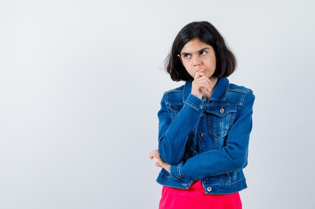 Chica joven de pie en pose de pensamiento en camiseta roja y chaqueta de jean y mirando pensativo, vista frontal.