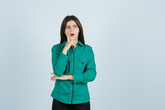 Chica joven de pie en pose de pensamiento, poniendo el dedo índice cerca de la boca en blusa verde, pantalón negro y mirando sorprendido. vista frontal.