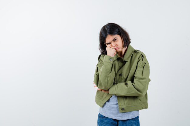 Chica joven de pie en pose de pensamiento en suéter gris, chaqueta de color caqui, pantalón de mezclilla y mirando pensativo. vista frontal.