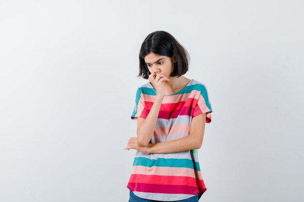 Chica joven de pie en posición de pensar, poniendo la mano en la boca en una camiseta a rayas de colores y mirando pensativa, vista frontal.