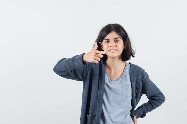 Chica joven poniendo una mano en la cintura, apuntando hacia la derecha con el dedo índice en una camiseta gris claro y una sudadera con cremallera frontal gris oscuro y luciendo linda.