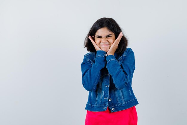 Chica joven poniendo las manos en las mejillas en camiseta roja y chaqueta de jean y mirando feliz, vista frontal.