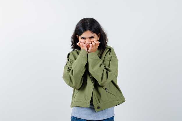 Chica joven poniendo las manos en las mejillas en suéter gris, chaqueta de color caqui, pantalón de mezclilla y mirando feliz. vista frontal.