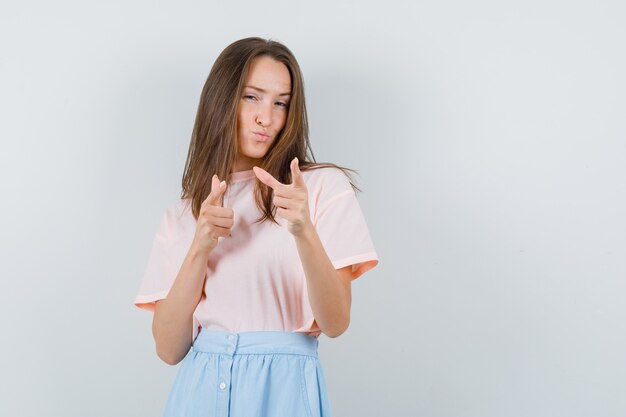 Chica joven posando con gesto de pistola en camiseta, falda y mirando confiado, vista frontal.