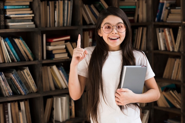 Chica joven positiva que sostiene un libro en la biblioteca