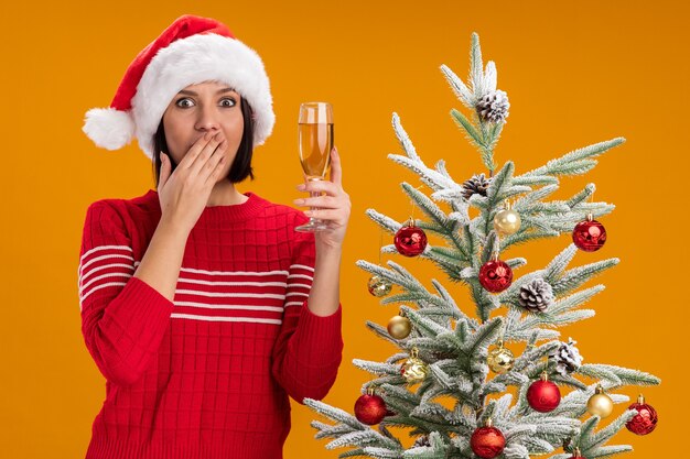 Chica joven preocupada con sombrero de santa de pie cerca del árbol de Navidad decorado sosteniendo una copa de champán manteniendo la mano en la boca aislada en la pared naranja