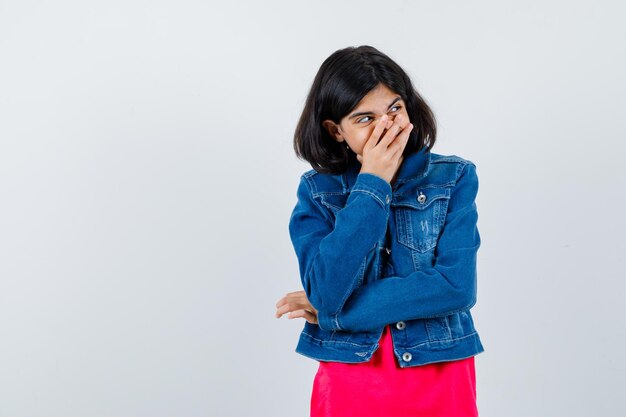 Chica joven que cubre la boca con la mano en camiseta roja y chaqueta de jean y se ve linda. vista frontal.