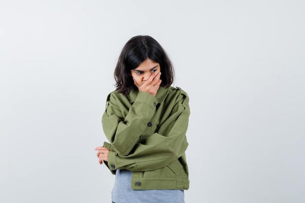 Chica joven que cubre la boca con la mano en suéter gris, chaqueta de color caqui, pantalón de jean y con aspecto cansado, vista frontal.