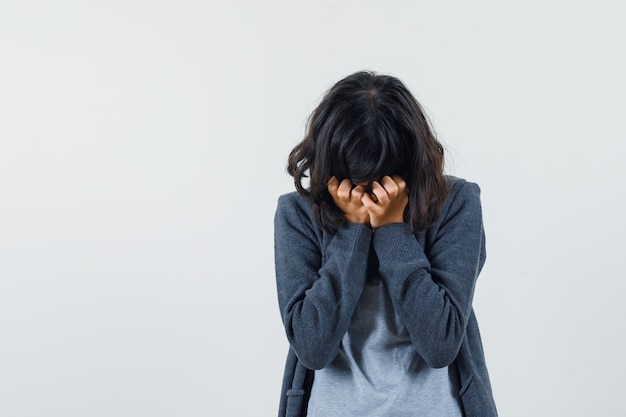 Chica joven que cubre la cara con la mano en una camiseta gris claro y una sudadera con capucha frontal con cremallera gris oscuro y parece estresada.