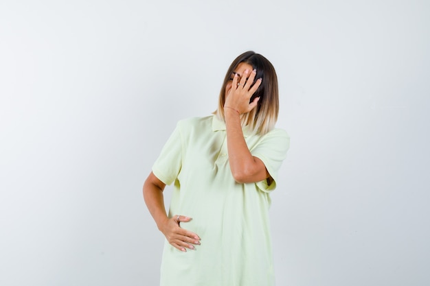 Chica joven que cubre la cara con la mano, sosteniendo la mano en el vientre en camiseta y luciendo exhausta. vista frontal.