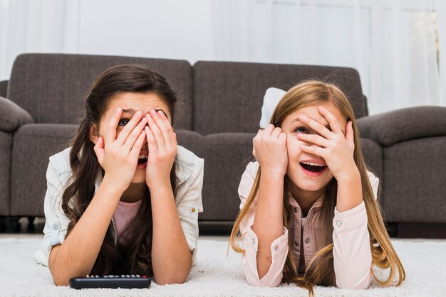 Chica joven que cubre la cara y mira a través de los dedos mientras ve la televisión en casa