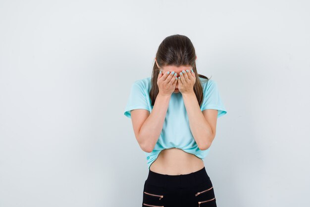 Chica joven que cubre la cara con las palmas en camiseta turquesa, pantalones y mirando molesto, vista frontal.