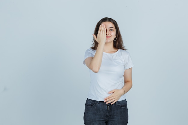 Chica joven que cubre el ojo con la mano en camiseta, jeans y mirando feliz, vista frontal.