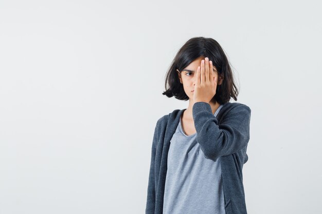 Chica joven que cubre parte de la cara con la mano en una camiseta gris claro y una sudadera con capucha gris oscuro con cremallera y se ve linda.