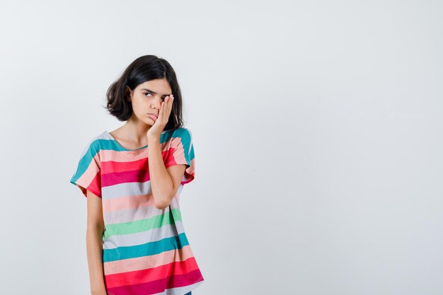 Chica joven que cubre parte de la cara con la mano en una camiseta a rayas de colores y parece agotada. vista frontal.