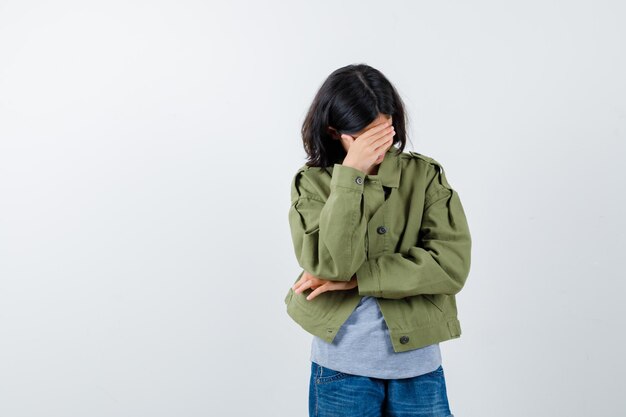 Chica joven que cubre parte de la cara con la mano en suéter gris, chaqueta de color caqui, pantalón de jean y con aspecto cansado, vista frontal.