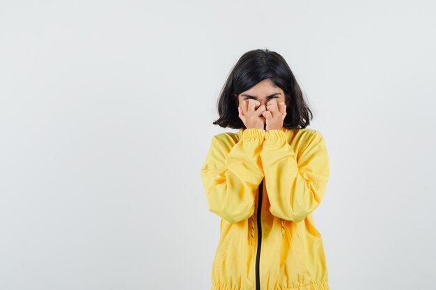 Chica joven que cubre parte de la cara con las manos en la chaqueta de bombardero amarilla y parece triste
