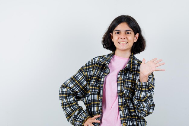 Chica joven que estira la mano como saludo a alguien mientras sostiene otra mano en la cintura con camisa a cuadros y camiseta rosa y parece amable. vista frontal.