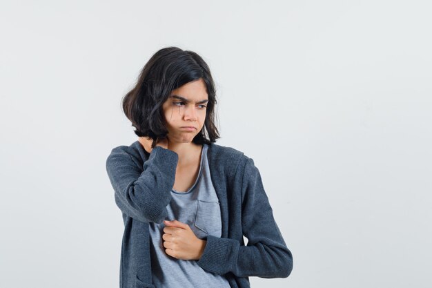 Chica joven que estira una mano mientras sostiene algo y lo señala con el dedo índice en una camiseta gris claro y una sudadera con capucha gris oscuro con cremallera y parece agotada