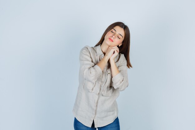 Chica joven que se inclina la mejilla en las manos en camisa beige, jeans y mirando soñoliento, vista frontal.