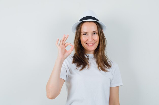 Chica joven que muestra un gesto aceptable en camiseta blanca, sombrero y mirando contento. vista frontal.