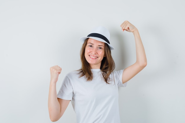 Chica joven que muestra el gesto del ganador en el sombrero de camiseta blanca y parece afortunado