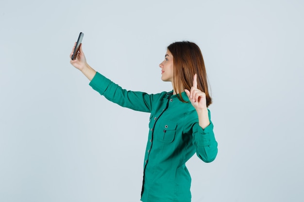 Chica joven que muestra el gesto de la pistola mientras hace una videollamada en blusa verde, pantalón negro y se ve linda, vista frontal.