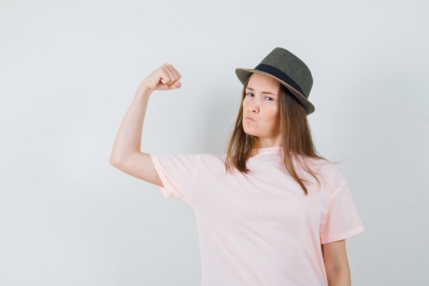 Chica joven que muestra los músculos del brazo en camiseta rosa, sombrero y parece seguro. vista frontal.