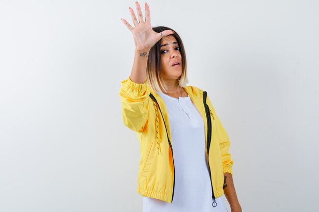 Chica joven que muestra la señal de pare en camiseta blanca, chaqueta amarilla y mirando confiado, vista frontal.