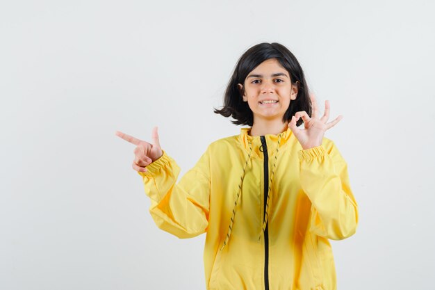 Chica joven que muestra el signo de ok y apunta a la izquierda con el dedo índice en una chaqueta de bombardero amarilla y parece optimista.