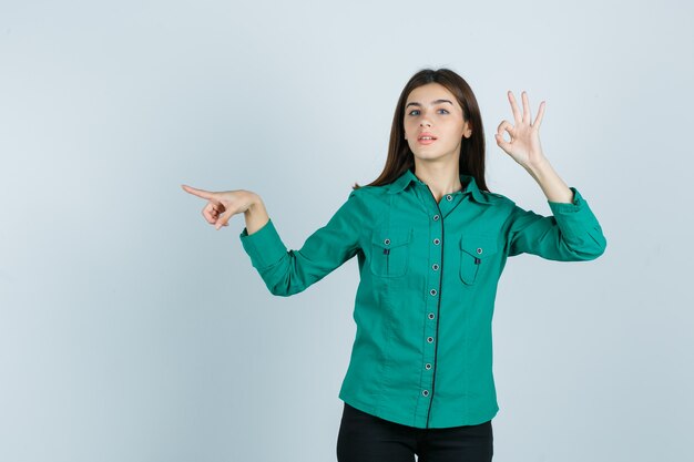 Chica joven que muestra el signo de ok, apuntando hacia la izquierda con el dedo índice en blusa verde, pantalón negro y mirando confiado, vista frontal