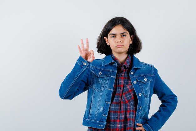 Chica joven que muestra el signo de ok mientras pone la mano en la cintura en camisa a cuadros y chaqueta de jean y mira seria, vista frontal.