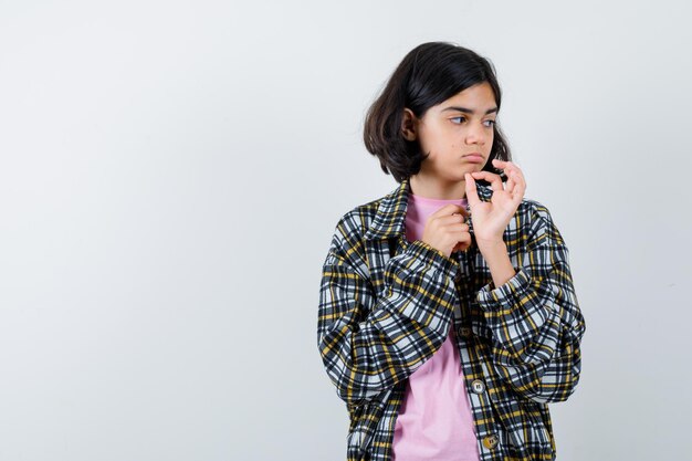 Chica joven que muestra el signo de ok, mirando a otro lado con camisa a cuadros y camiseta rosa y luciendo bonita, vista frontal.