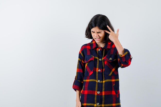 Chica joven que muestra el signo de la paz cerca del ojo, cerrando los ojos con camisa a cuadros y luciendo exhausta. vista frontal.