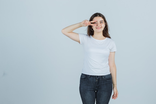 Chica joven que muestra el signo de la victoria en el ojo en camiseta, jeans y mirando dichoso, vista frontal.