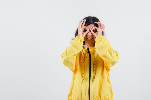 Chica joven que muestra signos bien en los ojos en chaqueta de bombardero amarilla y parece feliz