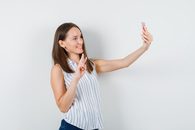 Chica joven que muestra v-sign mientras toma selfie en camiseta, jeans y parece feliz. vista frontal.