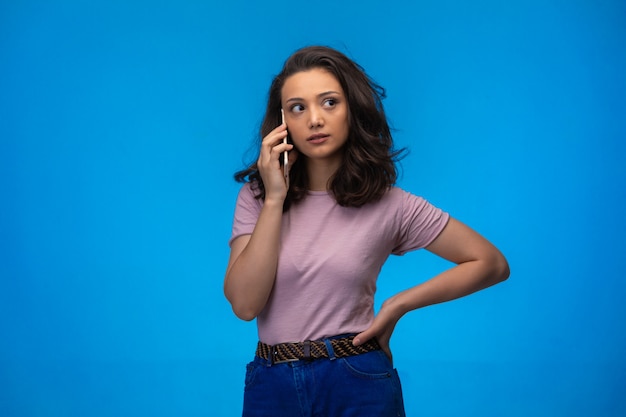 Foto gratuita chica joven que tiene una llamada en su teléfono inteligente mientras se lleva la mano a la cintura.