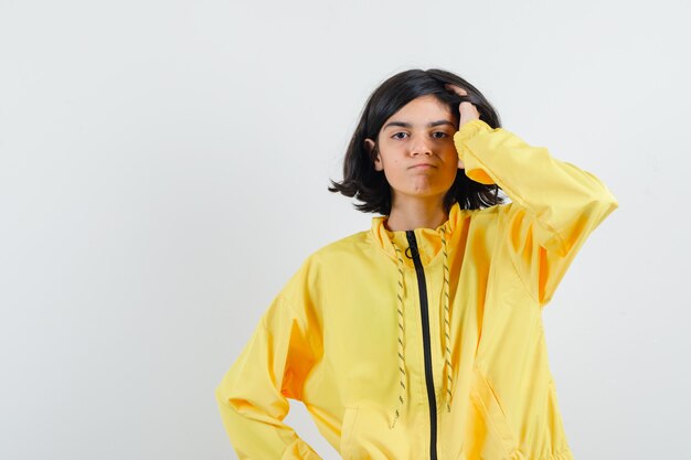 Chica joven rascándose la cabeza y sosteniendo una mano en la cintura en chaqueta de bombardero amarilla y mirando serio