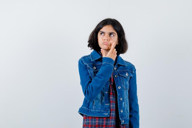 Chica joven rascándose la cara, mirando a otro lado con camisa a cuadros y chaqueta de mezclilla y mirando pensativo. vista frontal.