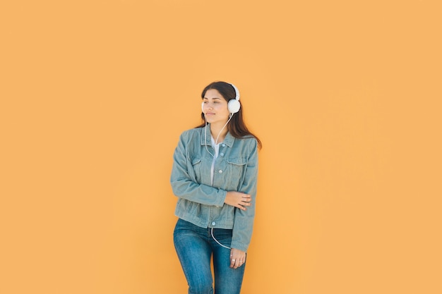 Chica joven relajante escuchando música con sus auriculares de pie frente a una pared amarilla