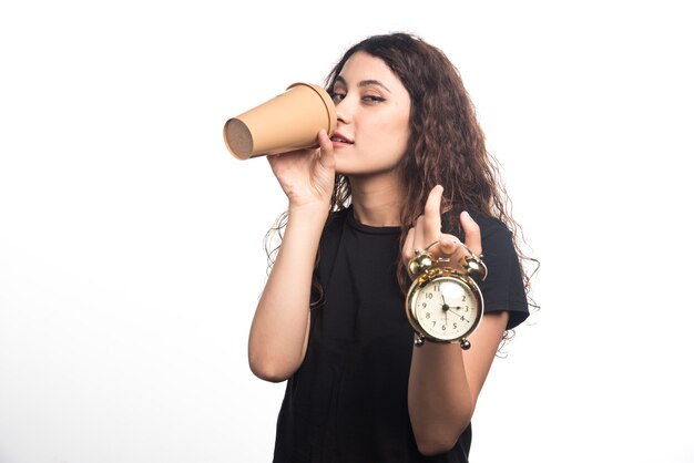 Chica joven con reloj en mano mostrando la hora y tomando café sobre fondo blanco. . Foto de alta calidad